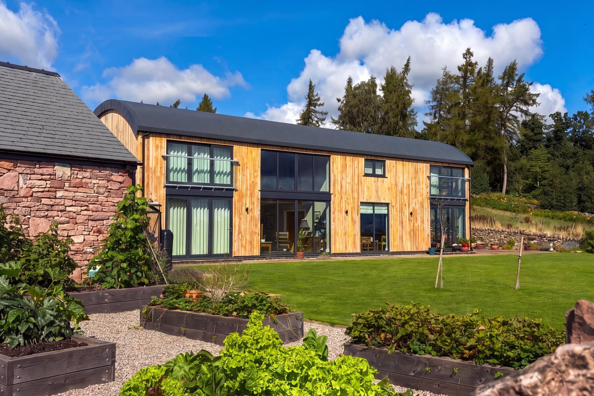 A completed Herdwick Barns home across its kitchen garden — larch cladding and full-height glazing under a curved slate roof, raised beds in the foreground