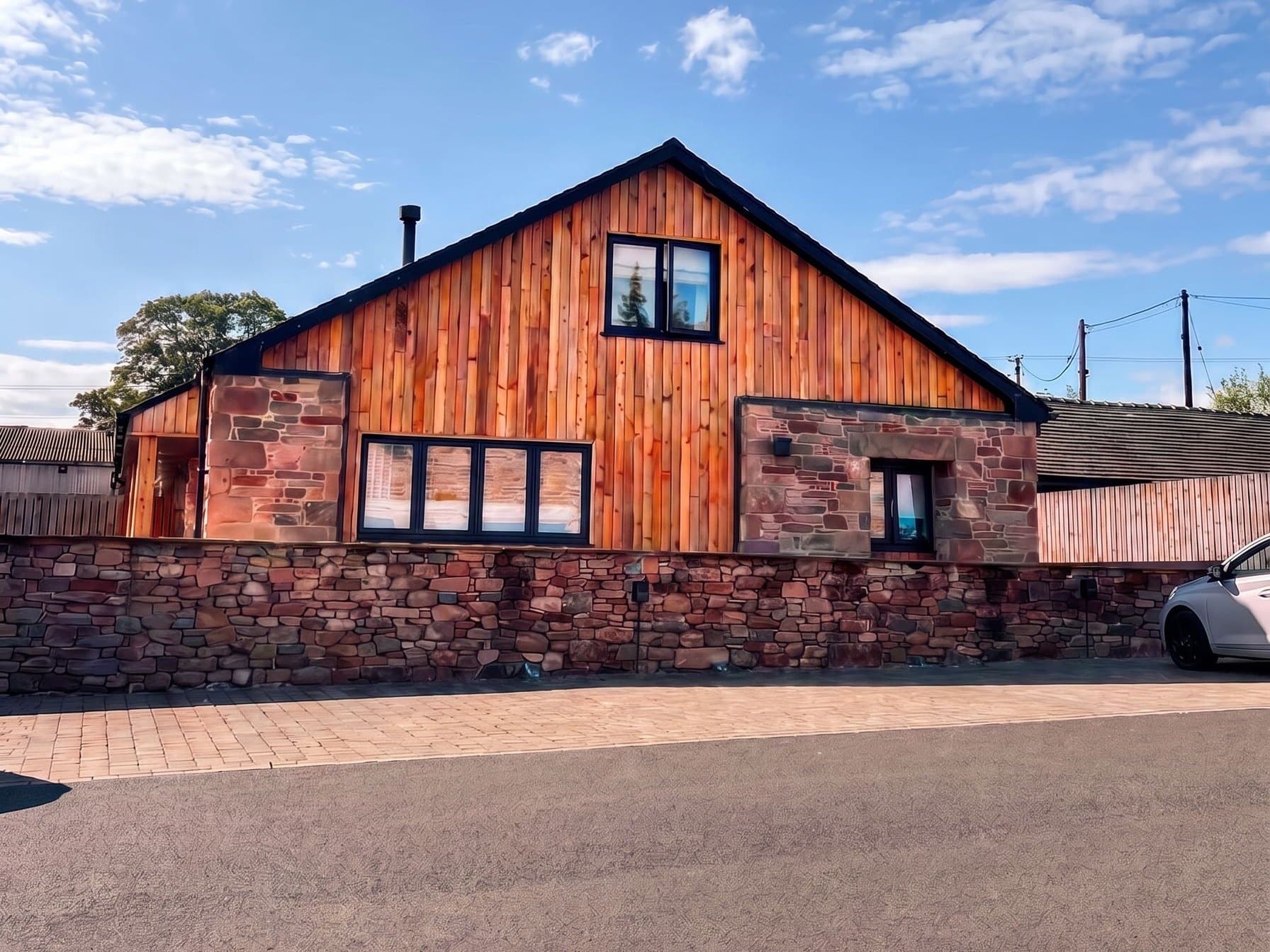 The gable of a Herdwick Barns home — larch cladding and red sandstone behind a dry-stone wall, under a clear blue sky