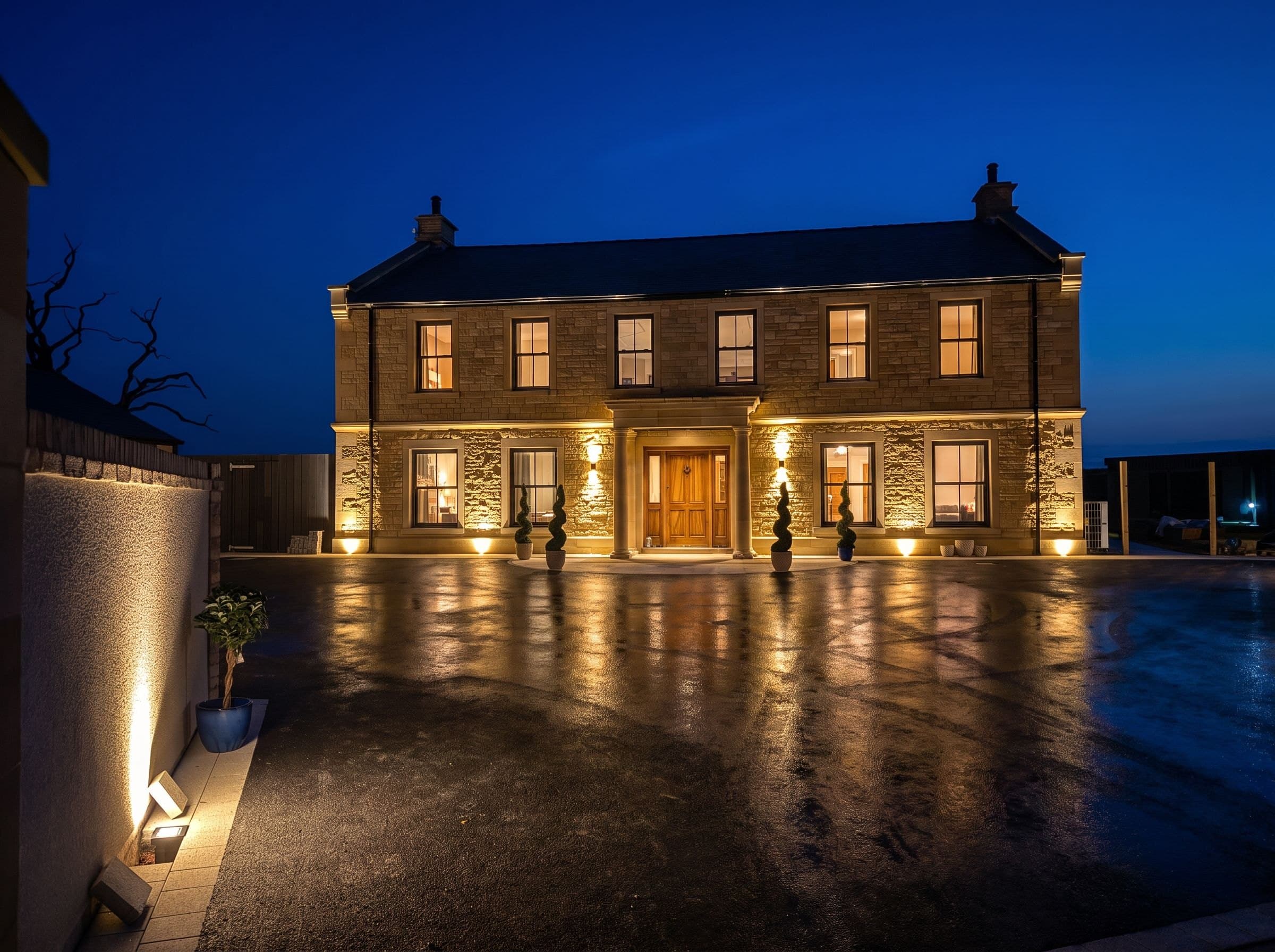 A grand Kiln Heights home in Lakeland stone at dusk — warm light from the sash windows and uplit facade reflected across the wet driveway