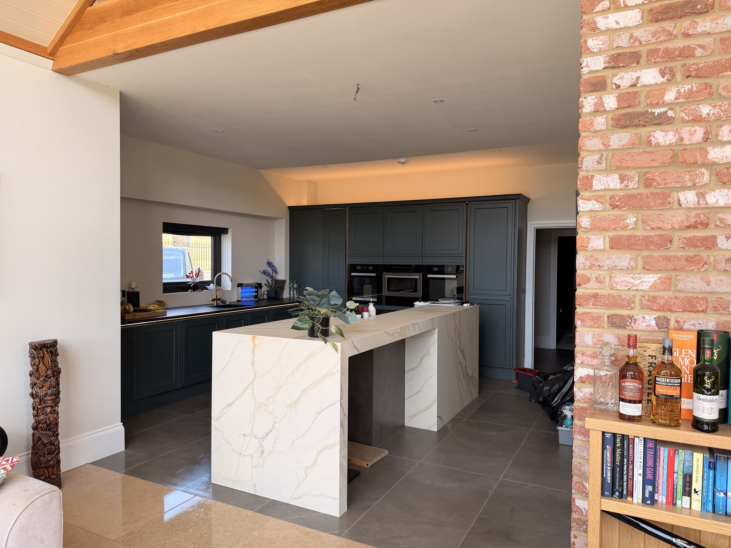 A kitchen at Swallows Rise — deep green cabinetry and a marble waterfall island, framed by an oak beam and reclaimed brick