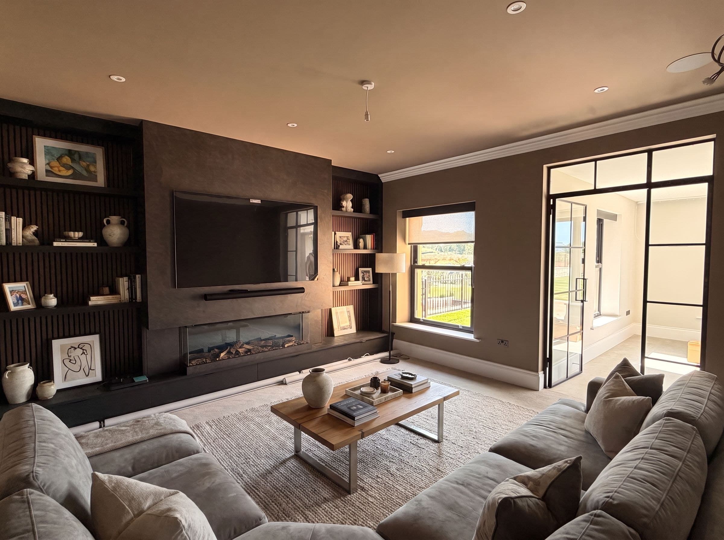 A furnished snug at Swallows Rise — deep sofas around a walnut media wall with a linear log-effect fire, beside a black-framed glazed screen